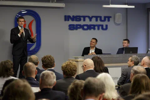 Wystąpienie prelegenta przy mikrofonie obok stołu prezydialnego na tle znaku Instytutu Sportu.