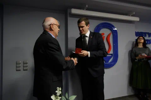 Wręczenie odznaczenia w czerwonym etui i uścisk dłoni na tle znaku Instytutu Sportu.
