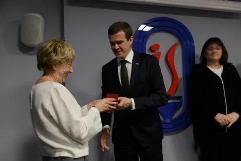Wręczenie odznaczenia w czerwonym etui wyróżnionej pracownicy w jasnym żakiecie.