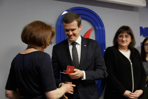 Przekazanie odznaczenia uczestniczce w ciemnej sukience na tle znaku Instytutu Sportu.