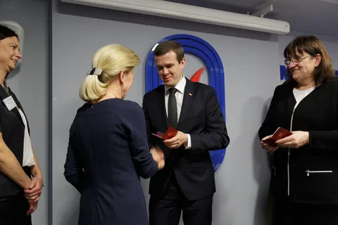Wręczenie odznaczenia uczestniczce w granatowej sukience na tle znaku Instytutu Sportu.