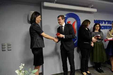 Uścisk dłoni podczas wręczania odznaczenia pracownicy w szarym kostiumie.
