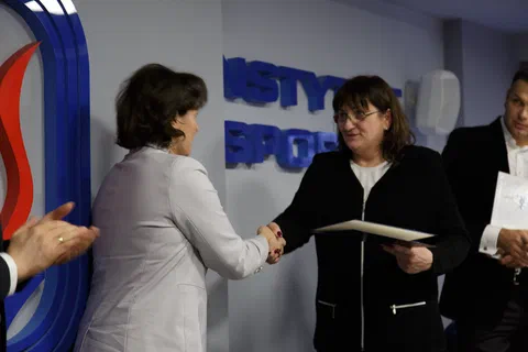 Wręczenie dyplomu i uścisk dłoni wyróżnionej pracownicy w jasnym żakiecie.