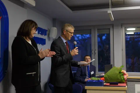Mężczyzna w garniturze odczytujący list gratulacyjny do mikrofonu.