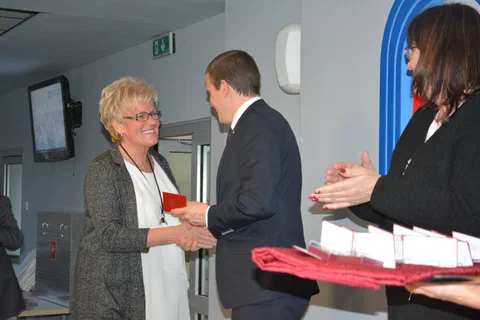 Przekazanie odznaczenia kobiecie w jasnej bluzce i szarym kardiganie.