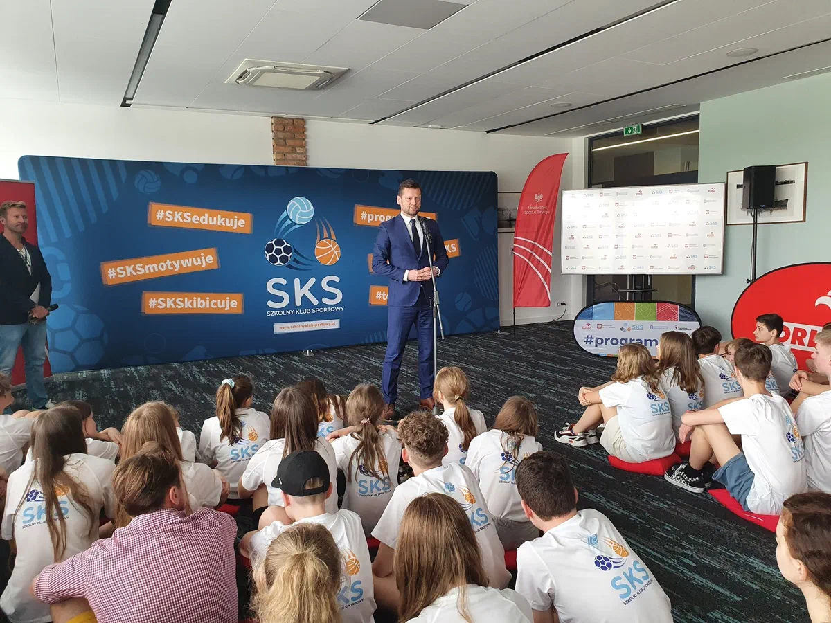 Spotkanie z dziećmi i młodzieżą w hali sportowej na tle ścianki programu Szkolny Klub Sportowy (#SKS).