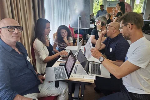 Praca grupowa uczestników przy stoliku z dwoma laptopami i arkuszem papieru.