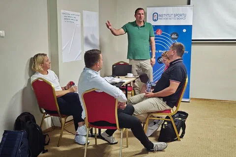 Uczestnik prezentujący plakaty na ścianie przed grupą siedzącą przy stoliku obok roll-upu Instytutu Sportu – PIB.