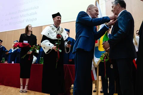 Przypinanie odznaczenia do marynarki wyróżnionego pracownika podczas uroczystości akademickiej w auli.