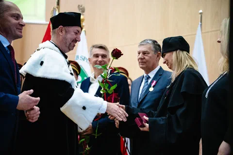 Rektor w todze i birecie wręczający czerwoną różę i składający gratulacje na tle pocztu sztandarowego.