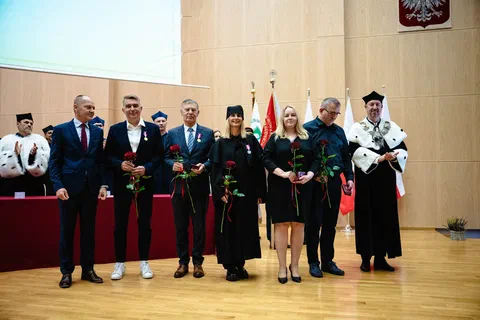 Wspólne zdjęcie odznaczonych osób z różami oraz władz uczelni w togach na tle pocztu sztandarowego i godła.