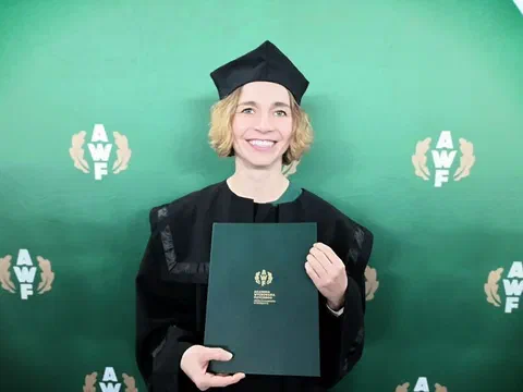 Dr hab. Dorota Sadowska w todze i birecie akademickim prezentuje dyplom habilitacyjny na tle ścianki AWF Warszawa