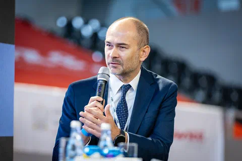 Prelegent w granatowym garniturze przemawiający do mikrofonu podczas debaty.
