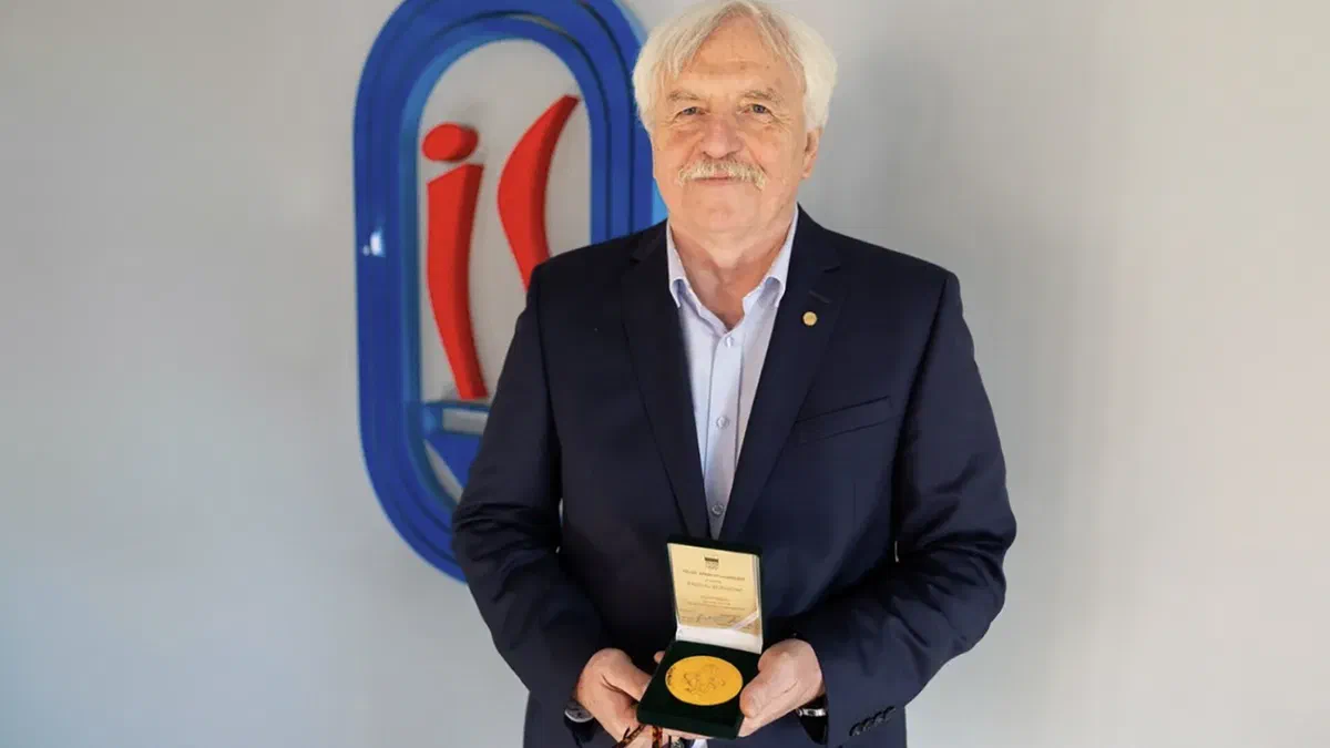 Dr Jerzy Eliasz prezentujący Złoty Medal za Zasługi dla Polskiego Ruchu Olimpijskiego w etui na tle znaku Instytutu Sportu – PIB.