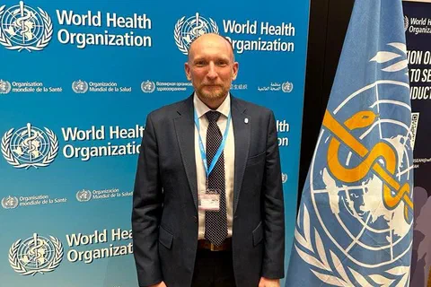 Uczestnik w garniturze stojący między ścianką i flagą Światowej Organizacji Zdrowia (WHO).
