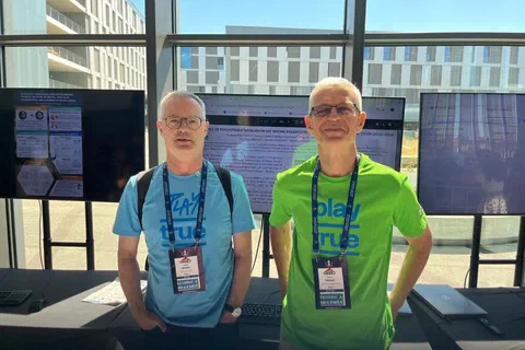 Dwaj badacze z identyfikatorami stojący przed monitorami podczas sesji e-posterowej.