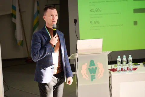 Prelegent w marynarce przemawiający przy mównicy na tle zielonego slajdu z wynikami procentowymi.