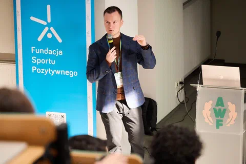 Prelegent z mikrofonem gestykulujący podczas wystąpienia obok baneru Fundacji Sportu Pozytywnego.