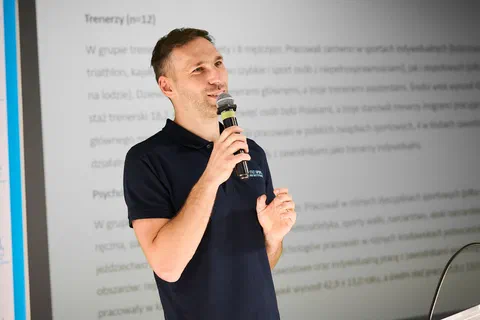 Zbliżenie na prelegenta z mikrofonem na tle slajdu opisującego badaną grupę trenerów i psychologów.