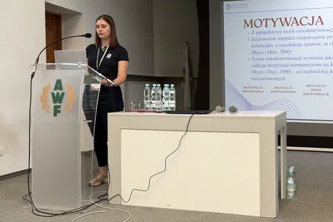 Prelegentka przy mównicy i stole prezydialnym obok ekranu ze slajdem poświęconym motywacji.