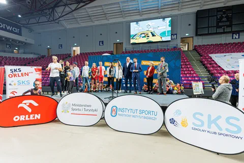 Grupa sportowców i organizatorów stojąca na scenie przed trybunami hali sportowej.