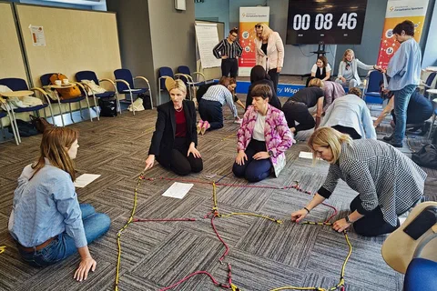 Uczestniczki budujące na dywanie figurę ze sznurków z widocznym w tle zegarem odliczającym czas zadania.