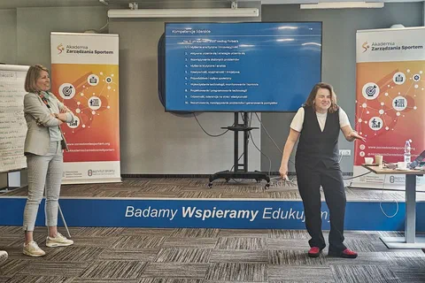 Dwie prowadzące omawiające slajd prezentacji o kompetencjach liderek przed ekranem i roll-upami Akademii Zarządzania Sportem.