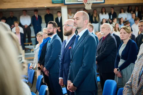 Zaproszeni goście w garniturach stojący w pierwszym rzędzie podczas części oficjalnej uroczystości.