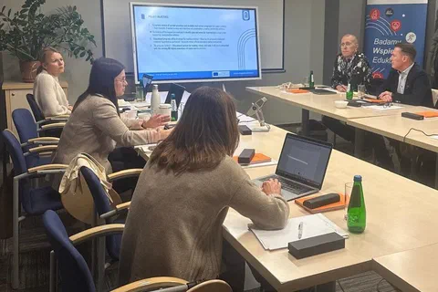 Uczestnicy spotkania projektowego siedzący przy stołach z laptopami podczas prezentacji przy ekranie.