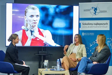 Trzy uczestniczki rozmowy siedzące w fotelach przed ekranem ze zdjęciem lekkoatletki i banerem Akademii Trenerskiej.