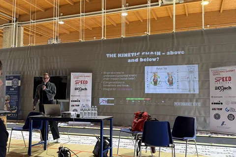 Prelegent występujący przy stoliku z laptopem przed ekranem projekcyjnym w hali szkoleniowej.