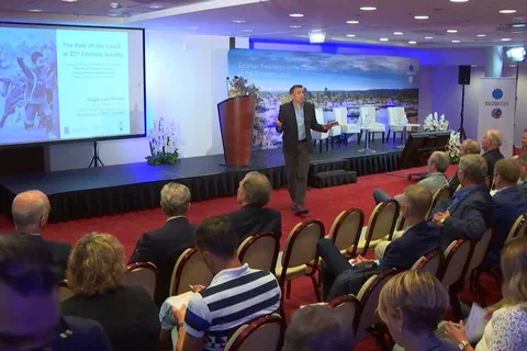 Prelegent przemawiający na scenie przed uczestnikami siedzącymi w pierwszych rzędach sali konferencyjnej.