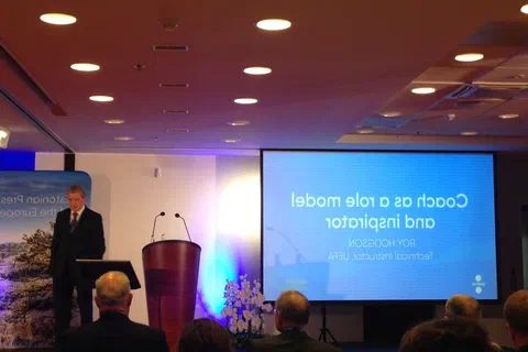 Mówca przy mównicy obok ekranu wyświetlającego pytanie o definicję trenera i coachingu.
