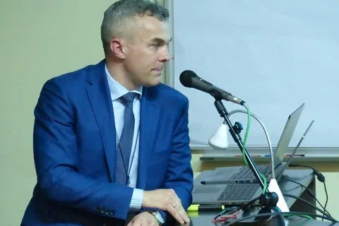 Prelegent w niebieskim garniturze przemawiający do mikrofonu przy stoliku z laptopem.