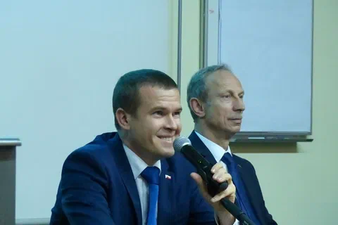 Dwaj mężczyźni w garniturach siedzący za stołem prezydialnym z mikrofonem.