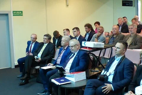 Grupa uczestników w garniturach siedząca w pobliżu projektora w sali wykładowej.