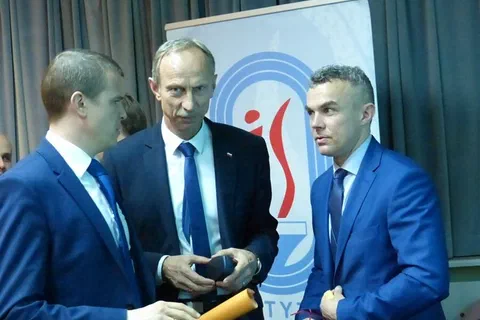 Trzej mężczyźni w garniturach rozmawiający na tle baneru Instytutu Sportu.