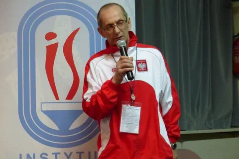 Prelegent w biało-czerwonym dresie reprezentacyjnym przemawiający do mikrofonu na tle baneru Instytutu Sportu.