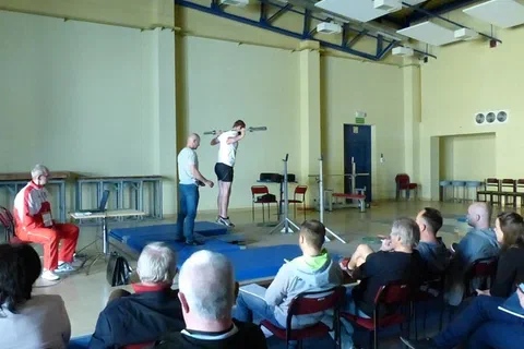 Pokaz ćwiczenia ze sztangą na pomoście przed grupą trenerów w sali sportowej.