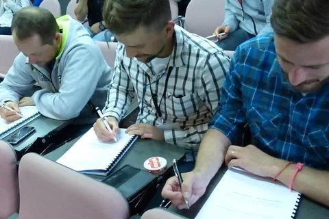 Trzej uczestnicy w koszulach w kratę i bluzie zapisujący notatki podczas wykładu.