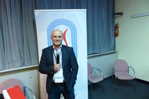 Prelegent w marynarce przemawiający przez mikrofon na tle baneru Instytutu Sportu.