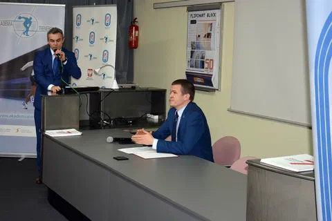 Wystąpienie otwierające przy stole prezydialnym na tle banerów Akademii Trenerskiej i Instytutu Sportu.