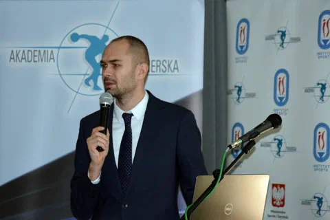 Prelegent w garniturze wygłaszający referat przy mikrofonie i laptopie.