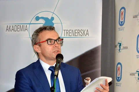 Wykładowca w okularach z notatkami w dłoni przemawiający do mikrofonu na tle ścianki Akademii Trenerskiej.