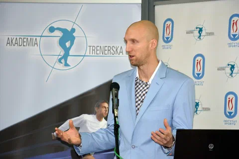 Prelegent w jasnej marynarce gestykulujący podczas wystąpienia przy mikrofonie.