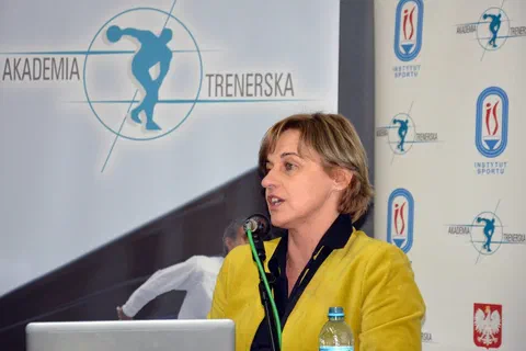 Kobieta w żółtym żakiecie przemawiająca do mikrofonu na tle ścianki Akademii Trenerskiej i Instytutu Sportu.