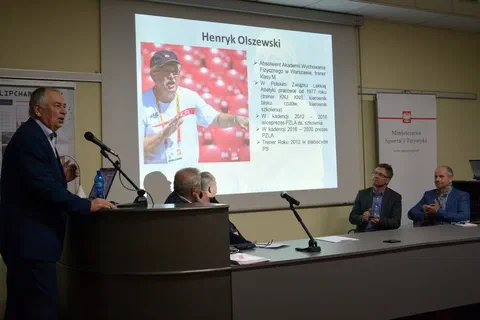 Uczestnicy panelu trenerskiego przy stole prezydialnym i mównicy pod ekranem projekcyjnym.