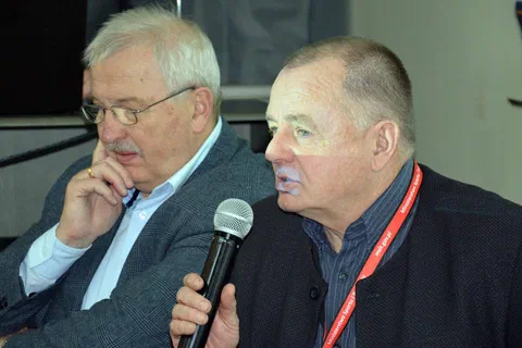 Zbliżenie na dwóch trenerów siedzących za stołem prezydialnym podczas wypowiedzi do mikrofonu.