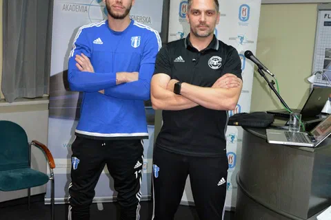 Dwaj mężczyźni w strojach sportowych stojący przy piłce gimnastycznej i banerach w sali.