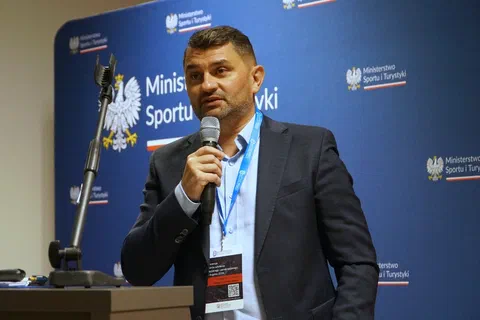 Mężczyzna w ciemnej marynarce przemawiający do mikrofonu na tle ścianki Ministerstwa Sportu i Turystyki.